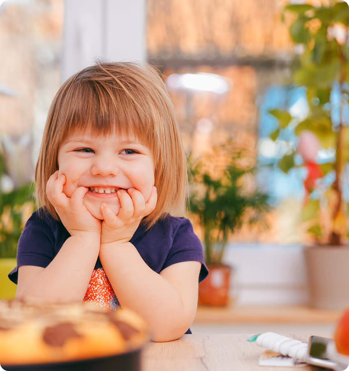 Ein lächelndes Kind, dass grinsend an einem Tisch in einer Kita-sitzt, bunter Hintergrund, bedeckter Tisch mit Spielzeug