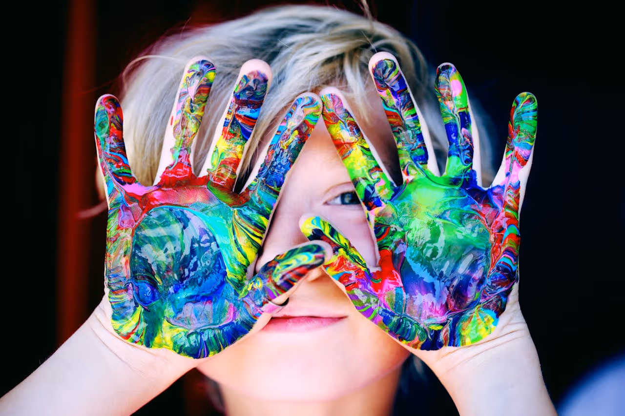 Kind, das bunt angemalte Handflächen in die Kamera hält und damit sein Gesicht verdeckt