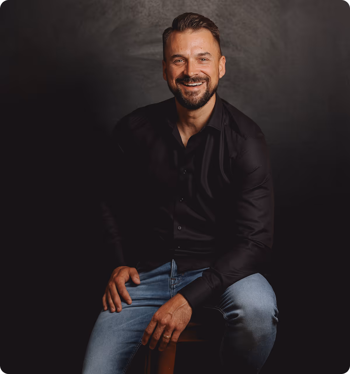Foto von Mario Behrens, dem purebo Gründer und Inhaber, der vor einem dunklen Hintergrund, auf einem Stuhl sitzend, lächelnd in die Kamera schaut.